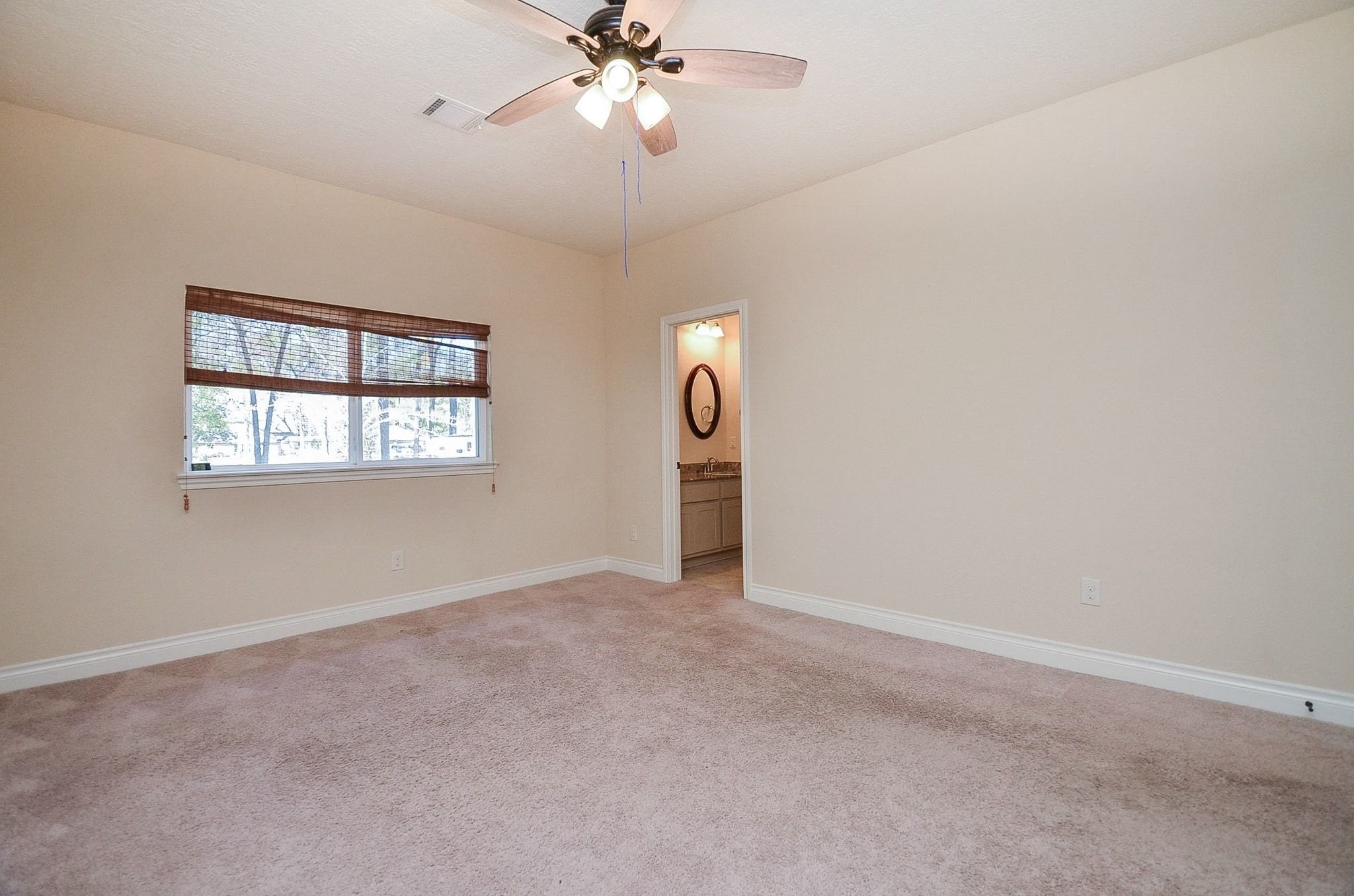 13327 Little Ranch Road Cypress, TX 77429 - Photo 29 of 49 This photo showcases a spacious bedroom with neutral walls and carpet, featuring a ceiling fan and natural light from a window with wooden blinds. It has an adjoining bathroom visible through an open doorway.
