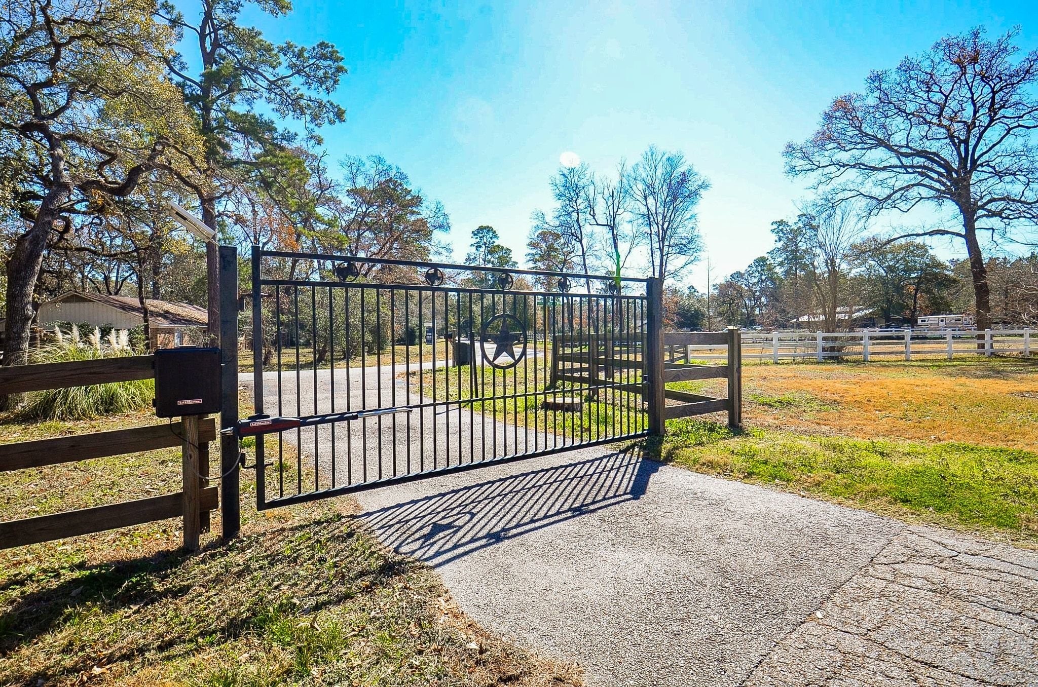 13327 Little Ranch Road Cypress, TX 77429 - Photo 8 of 49 This photo features a welcoming gated driveway entrance with a decorative metal gate and a paved path. It is surrounded by open land with mature trees, offering a sense of privacy and tranquility.