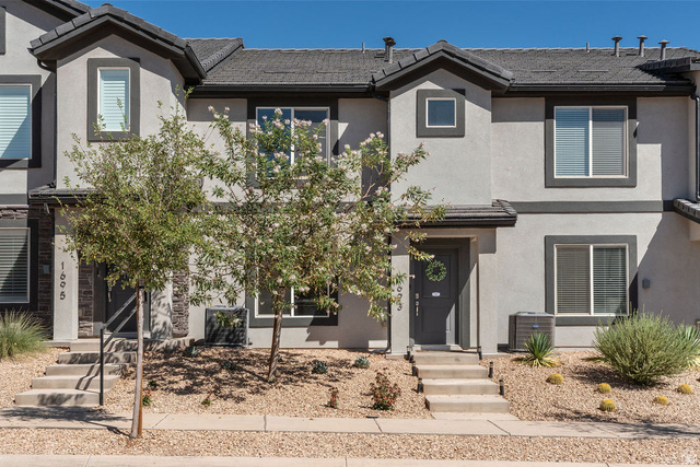 1693 Devils Gdn Lane Washington, UT 84780 - Photo 33 of 64 Gray stucco exterior with dark trim accents, featuring a dark gray front door, multiple windows with blinds, and mature desert landscaping