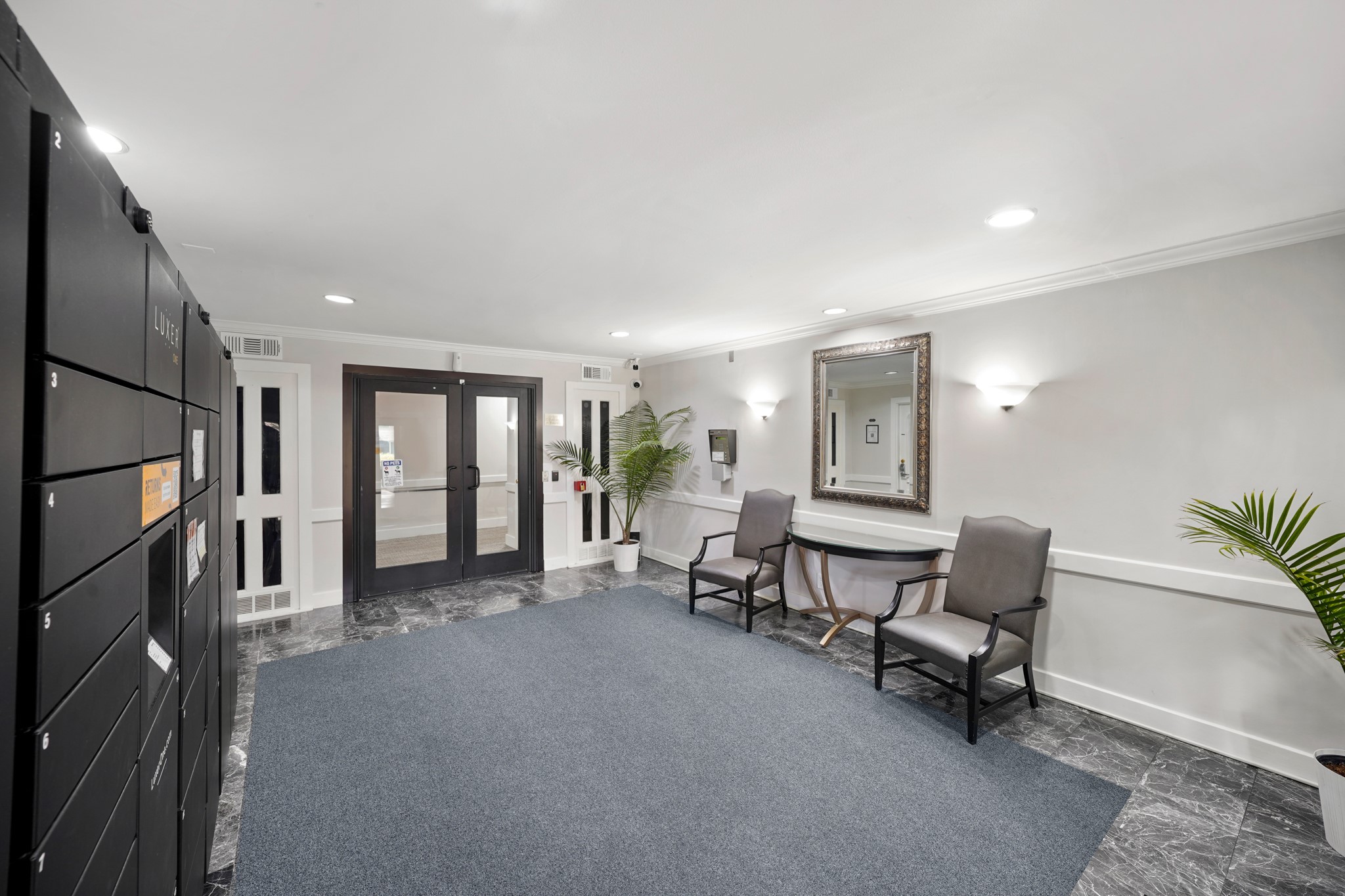 357 North Post Oak Lane, Unit 101 Houston, TX 77024 - Photo 12 of 19 A modern, cozy lobby featuring dark marble-style flooring, a large black smart-locker system, two sleek grey armchairs with a console table, and vibrant potted palm accents.