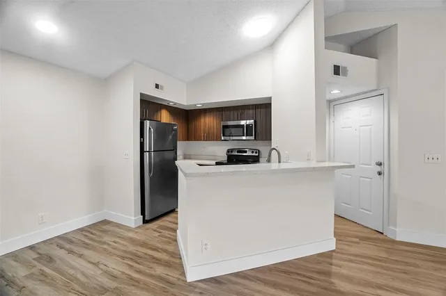 a living room with stainless steel appliances a wooden floor a large window and a kitchen view