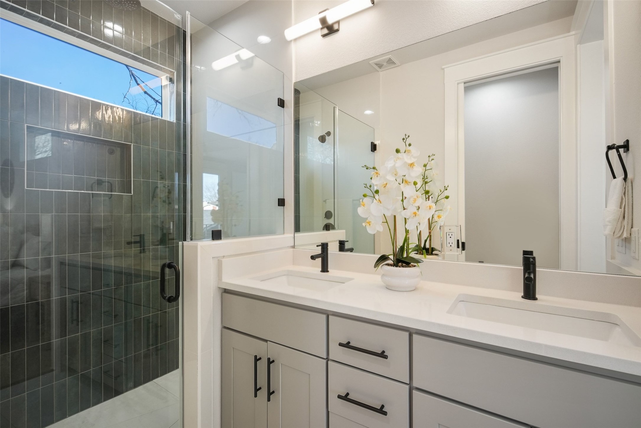 5109 Stone Gate Drive, Unit 2 Austin, TX 78721 - Photo 18 of 36 Contemporary bathroom featuring a dual vanity with light gray cabinetry and integrated sinks, matte black fixtures, a large mirror, and a glass-enclosed shower with vertical tile detailing and an integrated niche