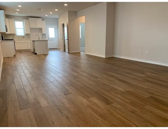 a view of an empty room and kitchen view with wooden floor