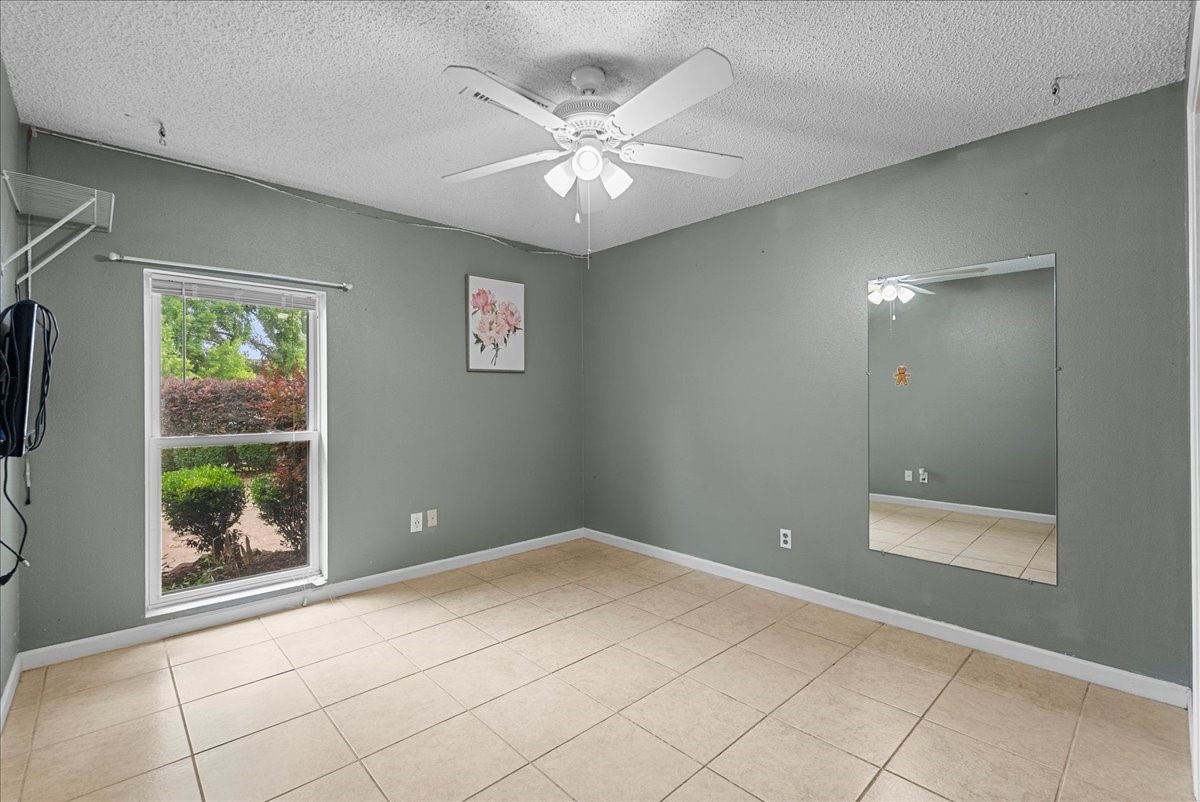 883 Silverpines Road Houston, TX 77062 - Photo 21 of 38 This bedroom features neutral gray walls, tile flooring, and a ceiling fan. It includes a large mirror, a window offering natural light with garden views, and a wall shelf.