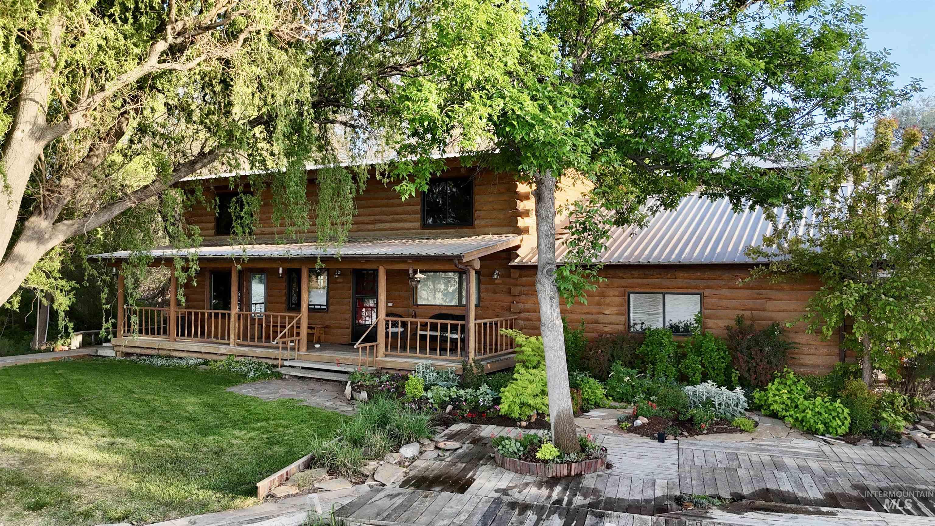 Log-sided residence featuring a full-length covered porch, metal roofing, mature trees, landscaped garden beds, and a wood-decked pathway
