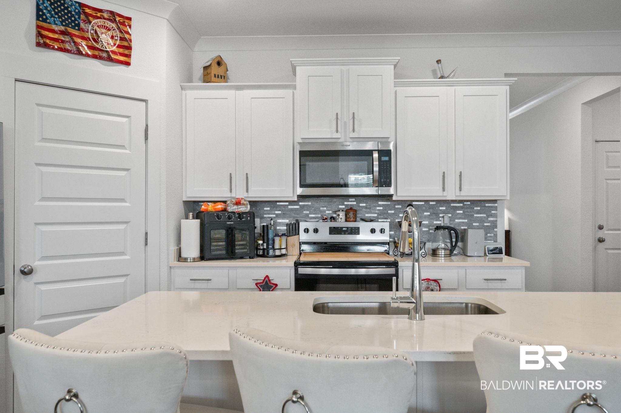 8508 Rosedown Lane Daphne, AL 36526 - Photo 11 of 29 Modern kitchen featuring a large island with an undermount sink and polished chrome faucet, white shaker-style cabinetry, stainless steel appliances, and a gray mosaic tile backsplash