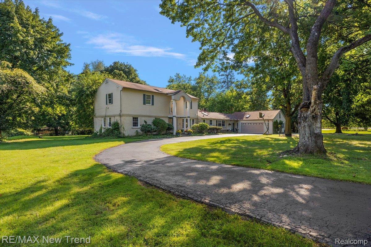 1450 Lenox Road Bloomfield Hills, MI 48304 - Photo 74 of 79 Expansive green lawn with mature trees and a paved driveway, featuring a two-story residence with light-colored siding, dark shutters, and an attached garage