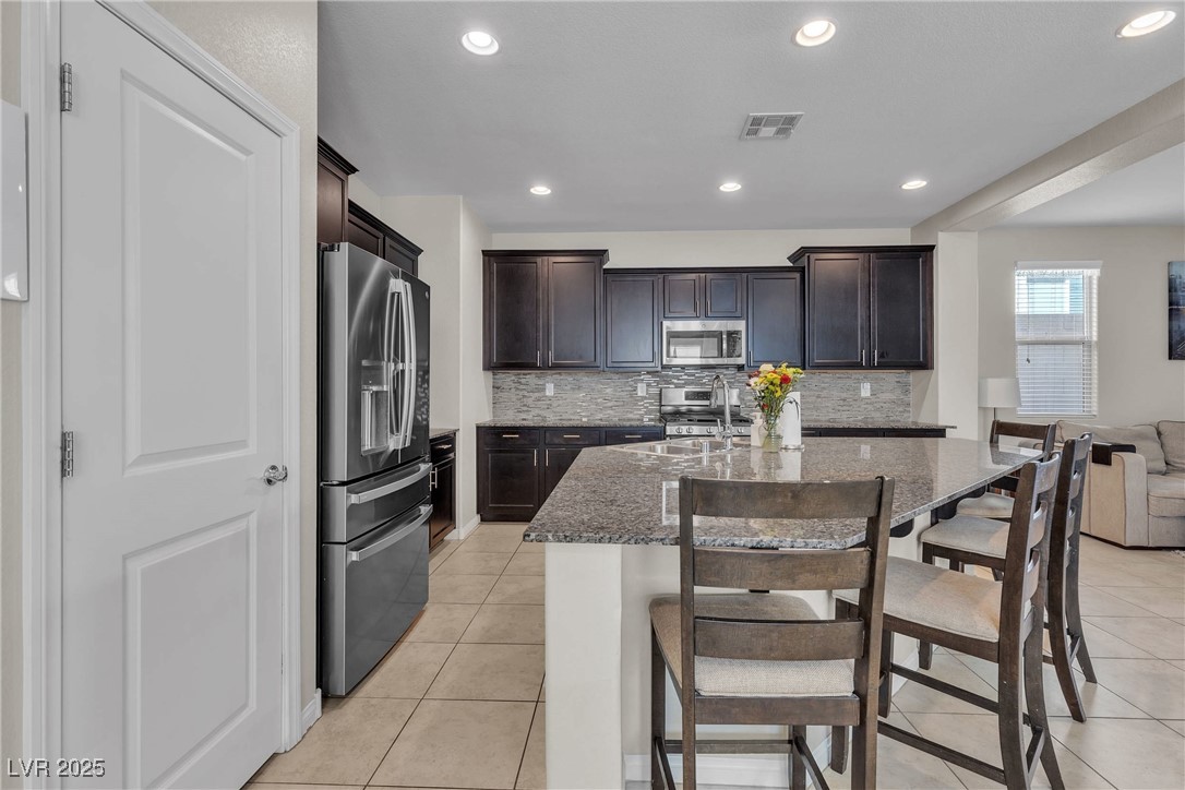 9950 Coyote Echo Court Las Vegas, NV 89166 - Photo 8 of 71 Kitchen with dark stone countertops, stainless steel appliances, a kitchen bar, dark brown cabinetry, and light tile patterned floors
