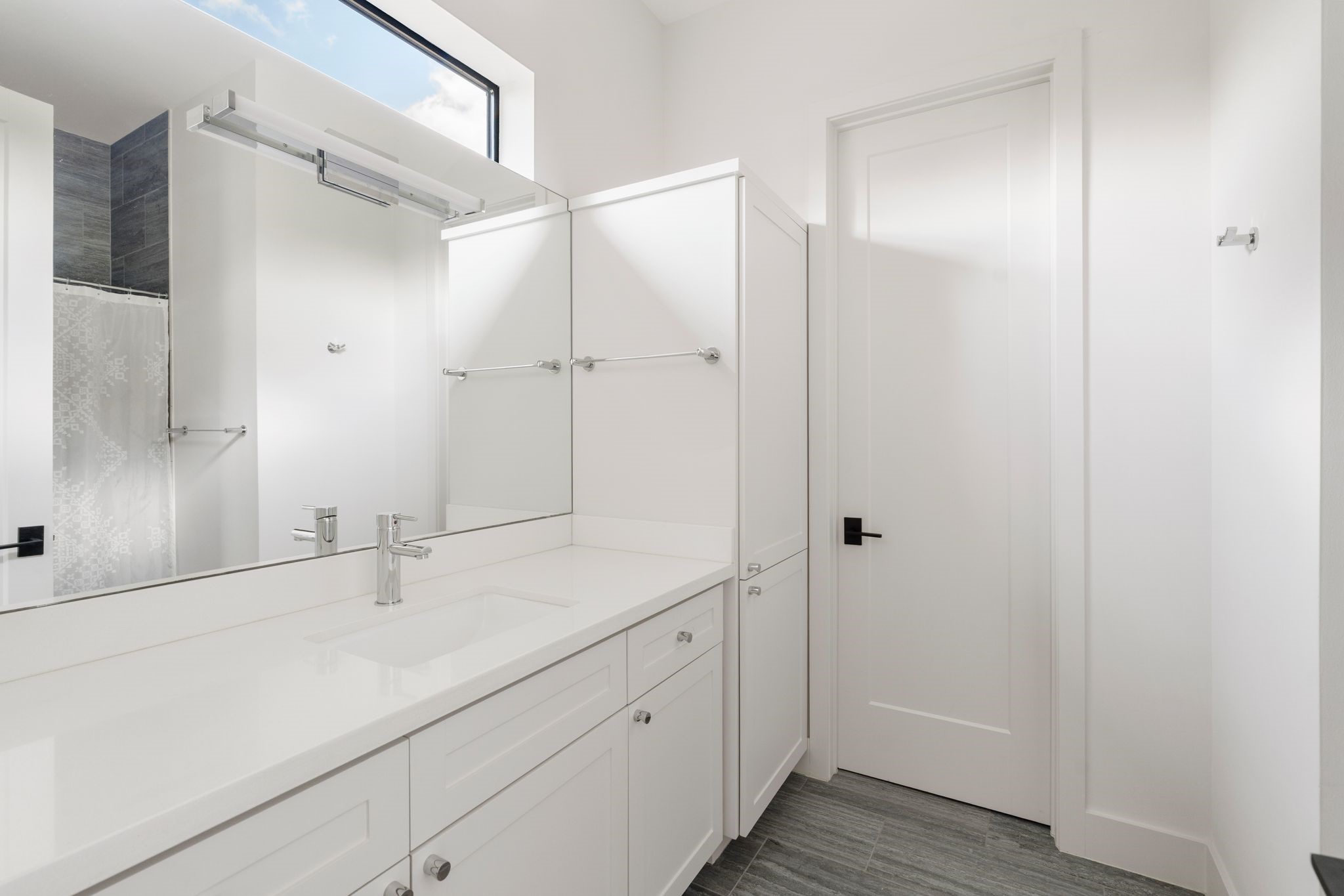 5427 Maple Street Houston, TX 77096 - Photo 33 of 46 This bathroom pairs a long vanity and oversized mirror with a tall built-in storage tower beside the door, with a tiled shower visible in the mirror's reflection.