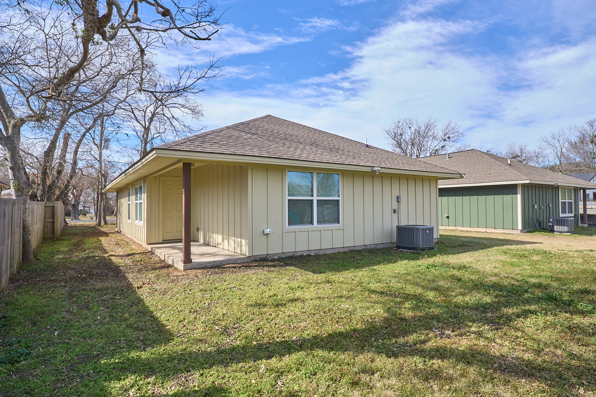1807 Nuches Lane Bryan, TX 77803 - Photo 23 of 27 This photo shows a single-story house with a light-colored exterior and a sloped roof. There's a small covered porch area and a fenced backyard, offering ample outdoor space. The surrounding area is open, with a few trees and neighboring houses.