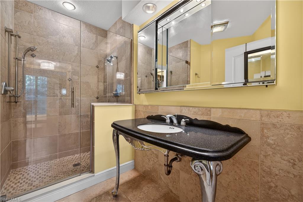 8420 Excalibur Circle, Unit R7 Naples, FL 34108 - Photo 18 of 24 Well-appointed bathroom featuring a glass-enclosed shower with tiled walls and floor, a vanity with a dark countertop and white basin, and a large mirror with integrated lighting