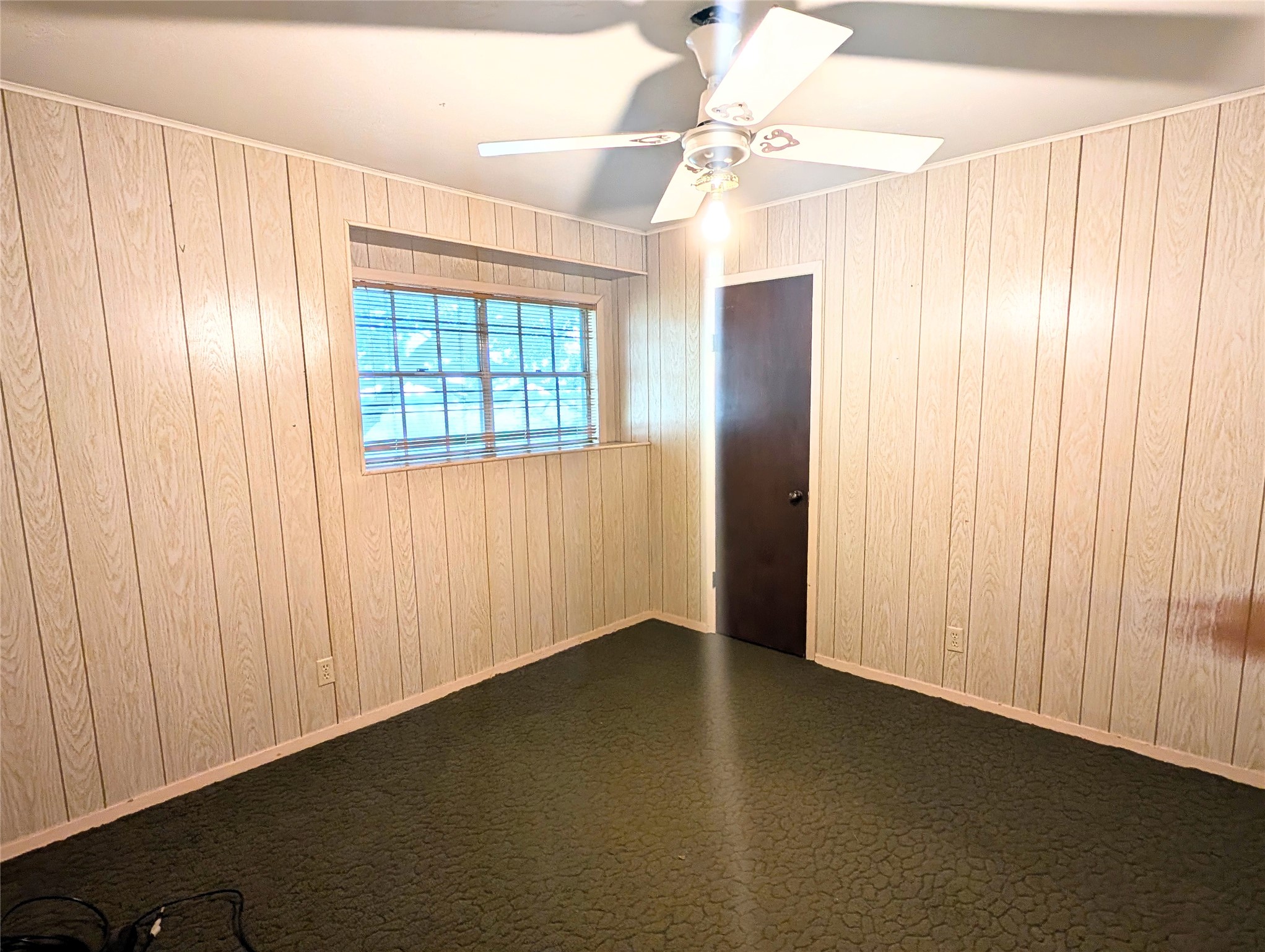 4779 Mixville Road Sealy, TX 77474 - Photo 26 of 47 Bedroom with wood panel walls, a ceiling fan, and a window for natural light. Features carpet and a dark wooden door, offering a classic and warm atmosphere.