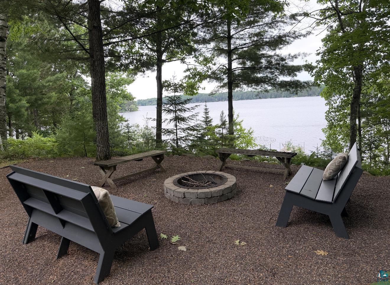 7338 South Maranatha Road Lake Nebagamon, WI 54849 - Photo 1 of 3 Lakefront fire pit area featuring a stone-ringed fire pit, gravel ground cover, mature trees, and multiple seating options
