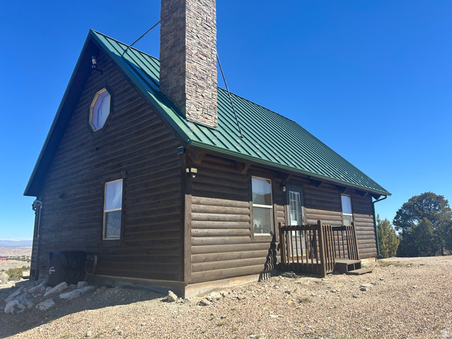 Log cabin exterior featuring a green metal roof, a stone-clad chimney, a small porch with wood railings, a unique octagonal window, and an exposed aggregate foundation