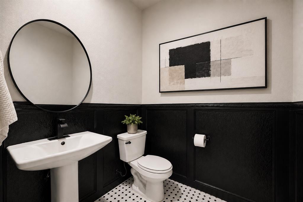 9828 Quail Pointe Road Aubrey, TX 76227 - Photo 12 of 23 Pedestal sink with a matte black faucet, round wall-mounted mirror, black wainscoting, white and black mosaic tile flooring, and a standard toilet