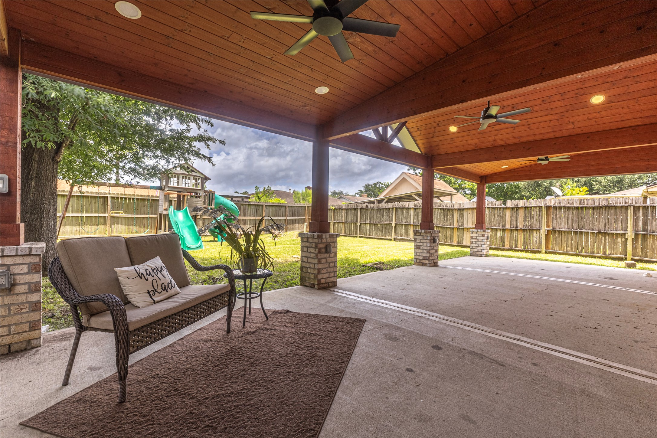 14419 Wadebridge Way Houston, TX 77015 - Photo 1 of 20 This photo showcases a spacious covered patio with wooden ceiling and fans, perfect for outdoor relaxation. It overlooks a fenced backyard with a playset, offering privacy space after a long day!