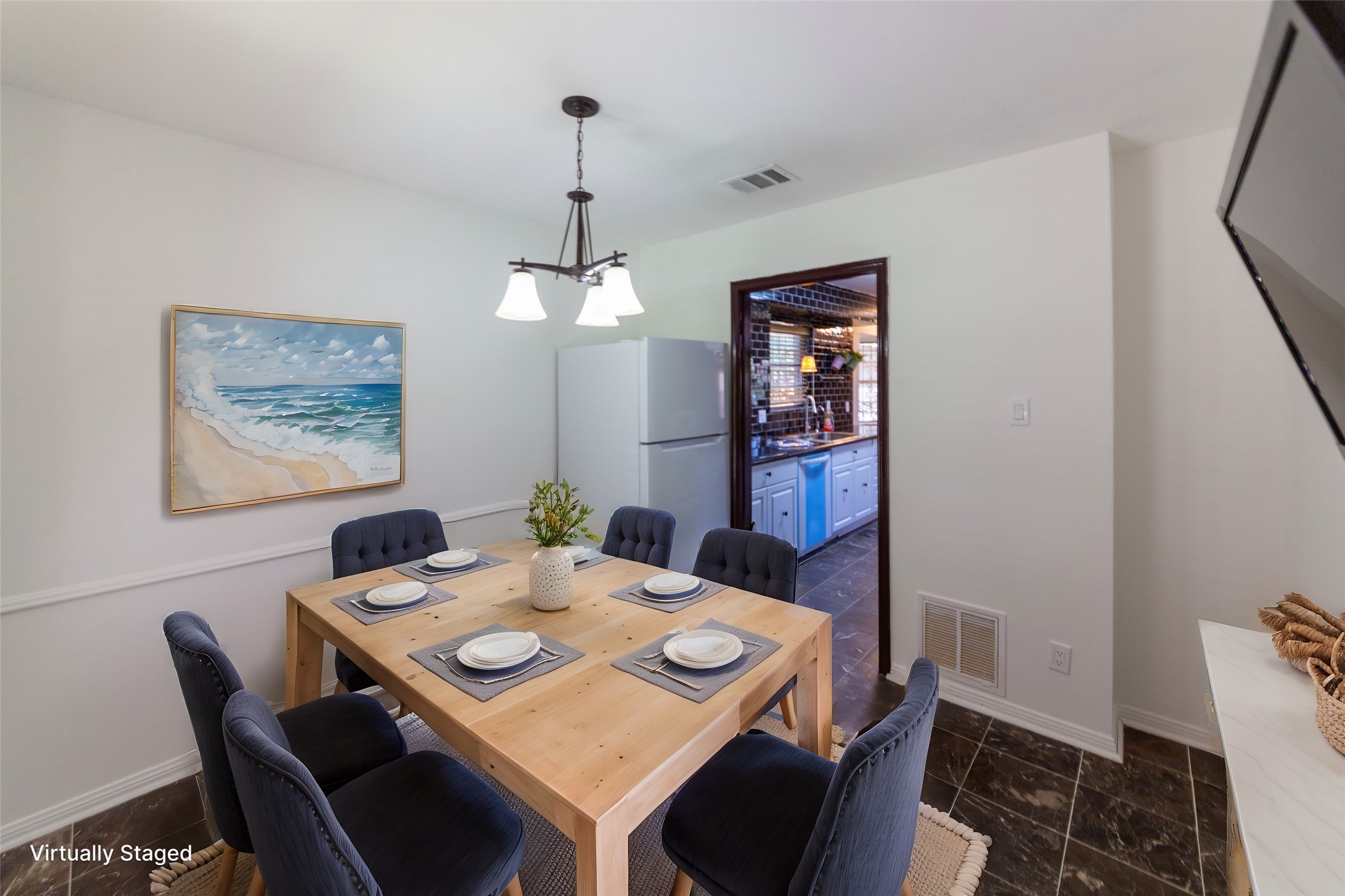 8826 Roos Road Houston, TX 77036 - Photo 12 of 42 A cozy dining area with a wooden table set for four, complemented by plush chairs. A calming beach painting adorns the wall. The room leads into a kitchen through an open doorway, revealing sleek cabinetry and a modern sink. The space is well-lit with a contemporary chandelier.