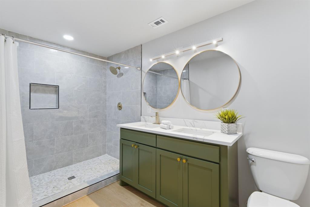 507 Country Club Road Cleburne, TX 76033 - Photo 26 of 39 Bathroom featuring a walk-in shower with gray tile surround and built-in niche, dual round mirrors with gold-tone frames, and a double vanity with an olive green finish and white countertop