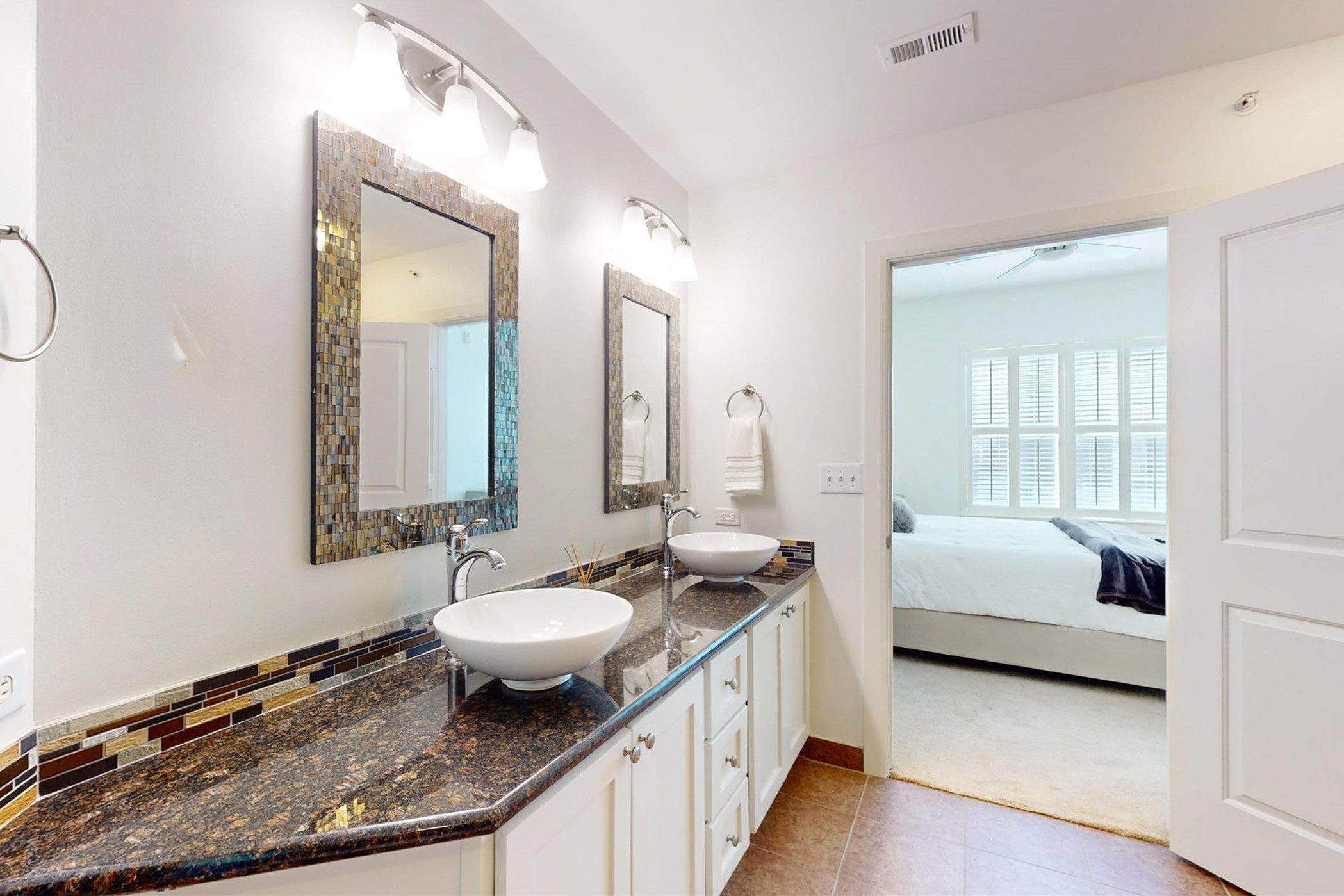 1010 Rosine Street, Unit 29 Houston, TX 77019 - Photo 9 of 16 A modern bathroom with dual vessel sinks on a granite countertop, featuring stylish tile backsplash and framed mirrors. The space is well-lit with overhead lighting and opens to a bright bedroom with large windows.