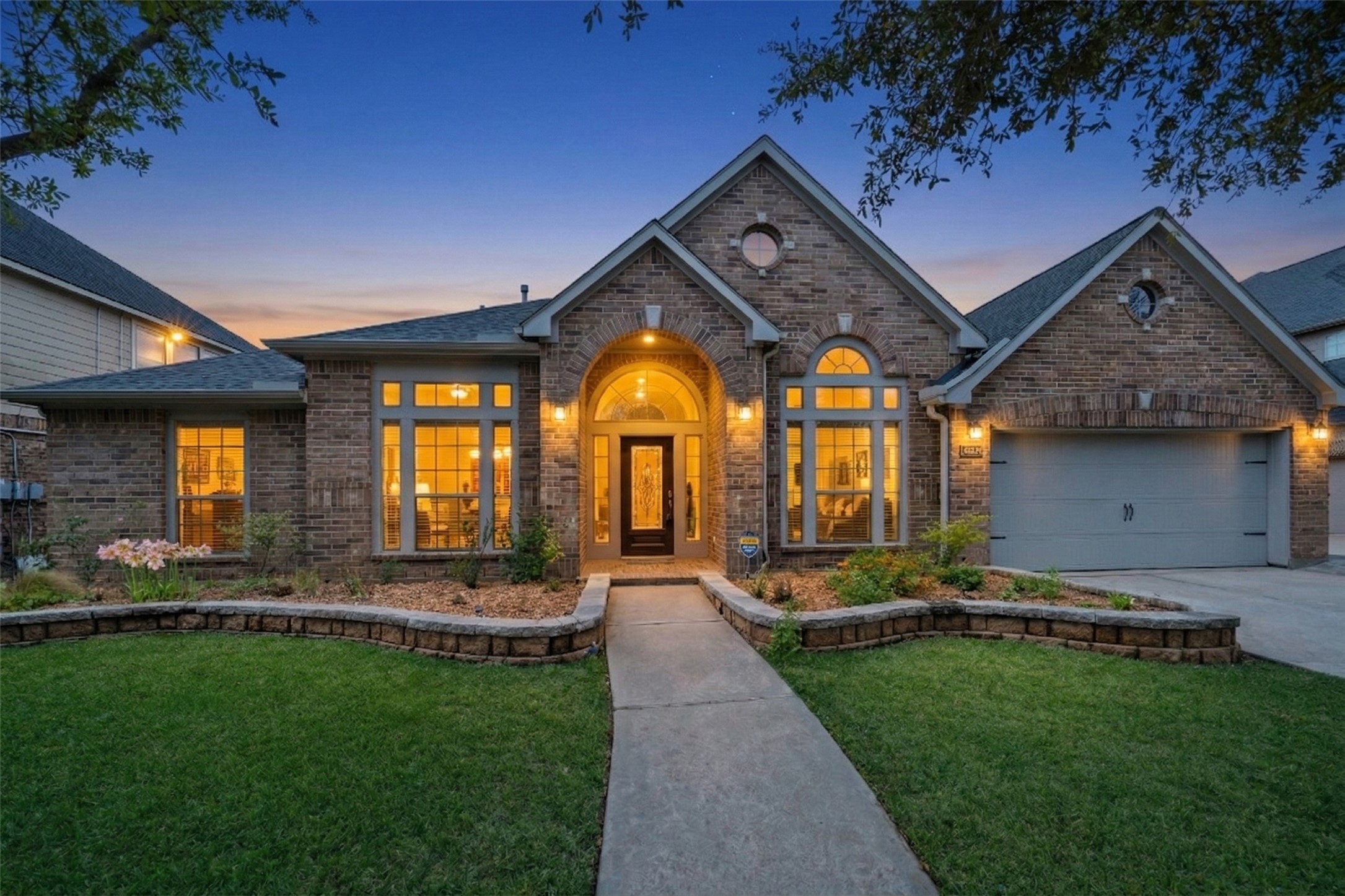 This inviting brick home features large, illuminated windows, a welcoming arched entryway, and a well-maintained raised flower beds. The three-car garage adds practicality to its charming curb appeal.