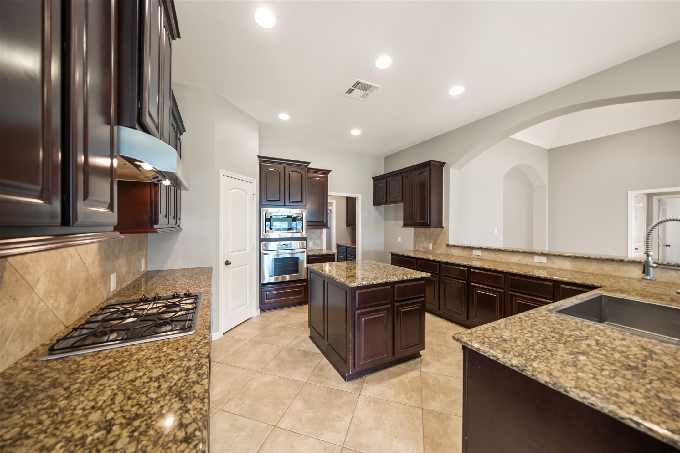 2818 Red Maple Drive Katy, TX 77494 - Photo 14 of 50 Spacious kitchen with elegant dark wood cabinets, granite countertops, and a central island. Features include a built-in oven, gas cooktop, and ample storage. Neutral tile flooring and recessed lighting create a bright, modern atmosphere.