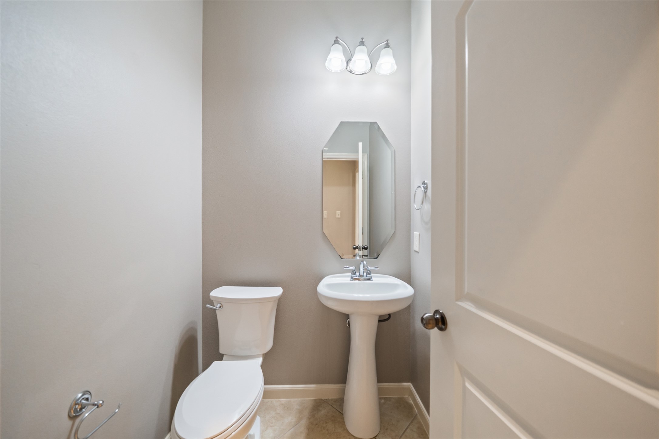 2818 Red Maple Drive Katy, TX 77494 - Photo 19 of 50 Convenient half-bathroom just off the entryway featuring a pedestal sink, toilet, and an octagonal mirror. The space is well-lit with a three-light fixture and has neutral-toned walls and tiles.