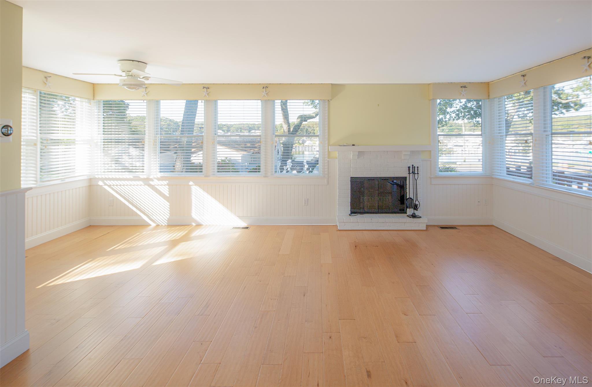 1 Canal Road Hampton Bays, NY 11946 - Photo 13 of 50 Unfurnished living room featuring a brick fireplace, light wood-type flooring, wainscoting, and ceiling fan