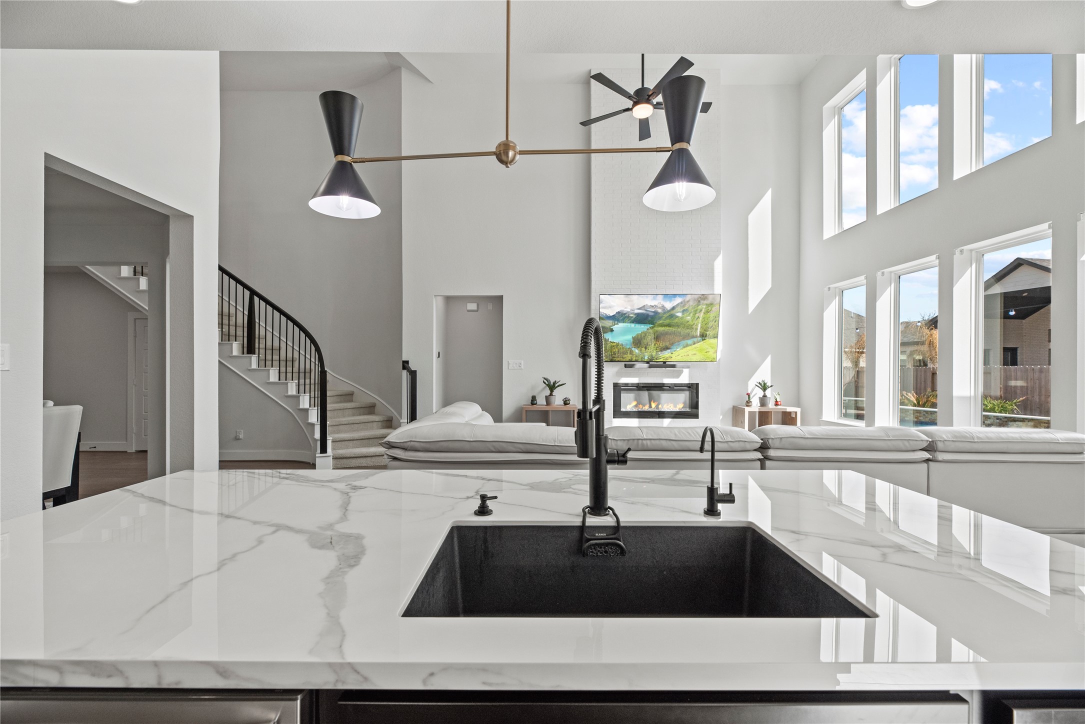 30155 GOLD FINCH Place Fulshear, TX 77441 - Photo 9 of 26 Stunning open-concept design highlighted by a dramatic two-story living room, floor-to-ceiling windows, and a sleek fireplace feature wall. The oversized quartz island with a modern undermount sink and statement lighting overlooks the living space—perfect for entertaining while enjoying views of the backyard and pool.