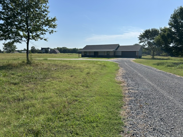 190 South 3 Mile Road Fort Gibson, OK 74434 - Photo 27 of 35 Gravel driveway leading to a single-story residence with a dark roof and light-colored siding, surrounded by expansive green space