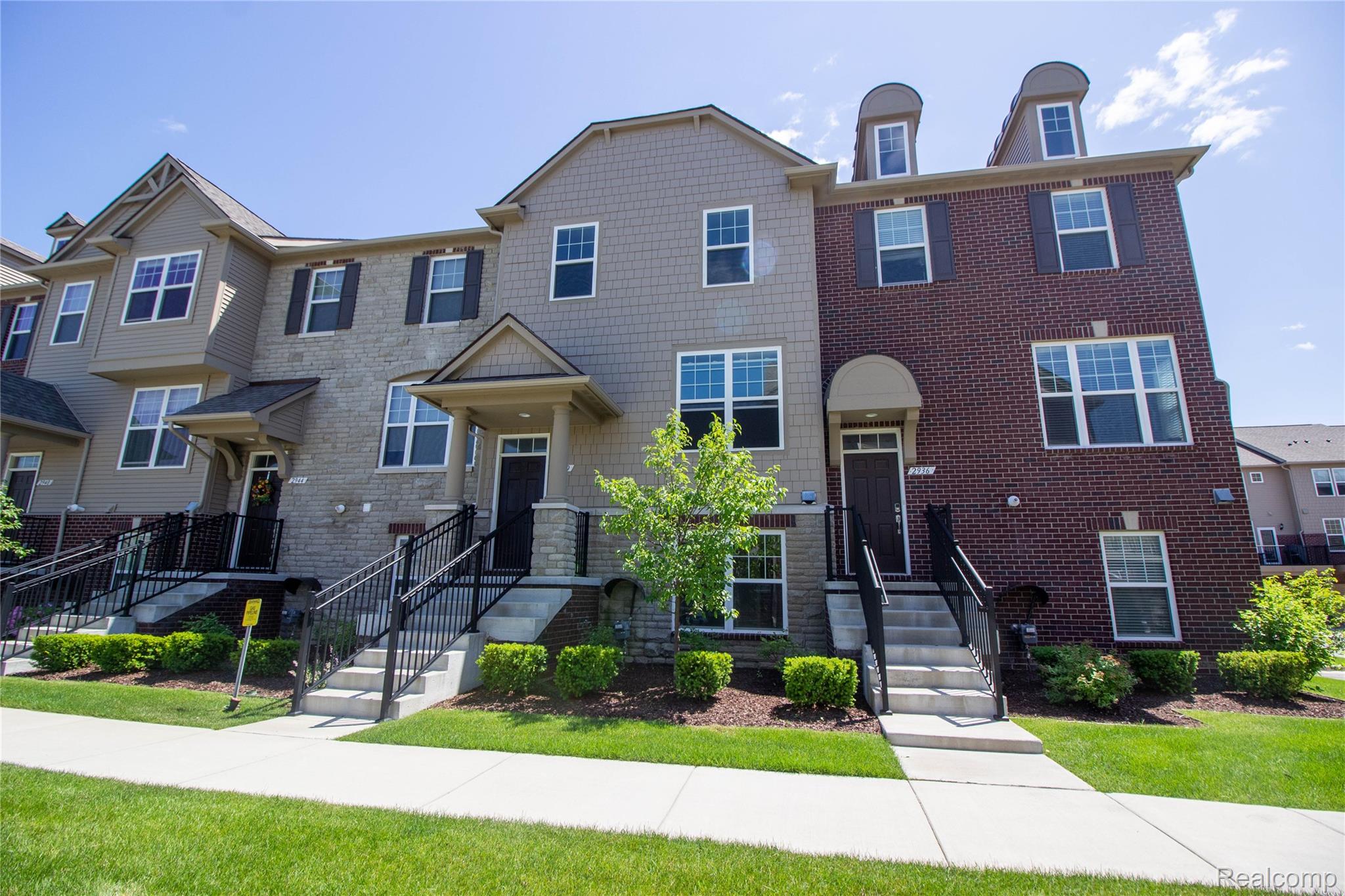 2940 Glenbar Circle Rochester, MI 48307 - Photo 1 of 15 Townhome exterior featuring varied siding and brick facades, individual covered entryways, concrete steps with black metal railings, manicured shrubbery, and prominent dormer windows