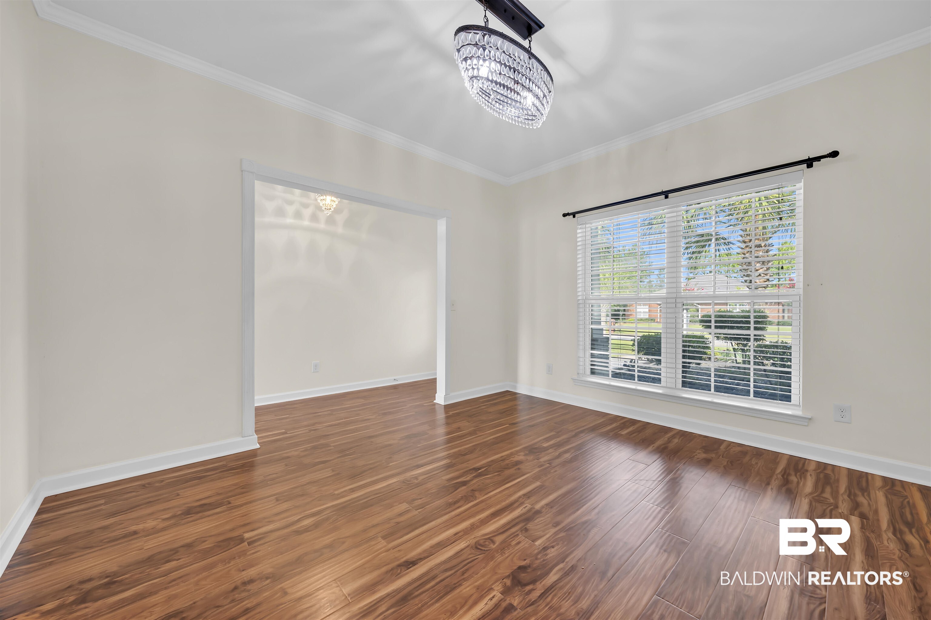 10147 Lyttleton Loop Lillian, AL 36549 - Photo 11 of 29 Spacious room featuring rich wood-finish flooring, a large window with blinds, an ornate chandelier, white crown molding, and a wide cased opening to an additional room