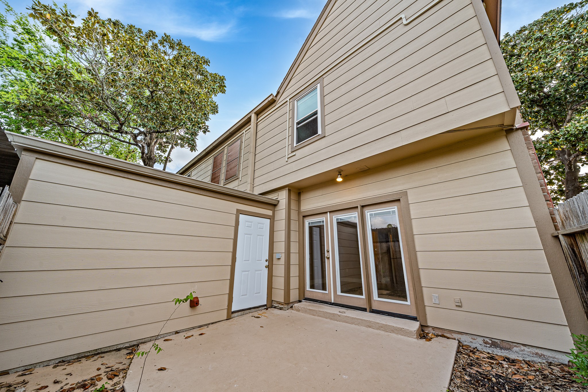 830 Threadneedle Street, Unit 240 Houston, TX 77079 - Photo 27 of 33 This photo shows a cozy backyard with a concrete patio, surrounded by a light-colored exterior of a two-story house. It features a set of French doors leading inside and a small storage area on the side. The space is enclosed with a wooden fence and shaded by nearby trees.
