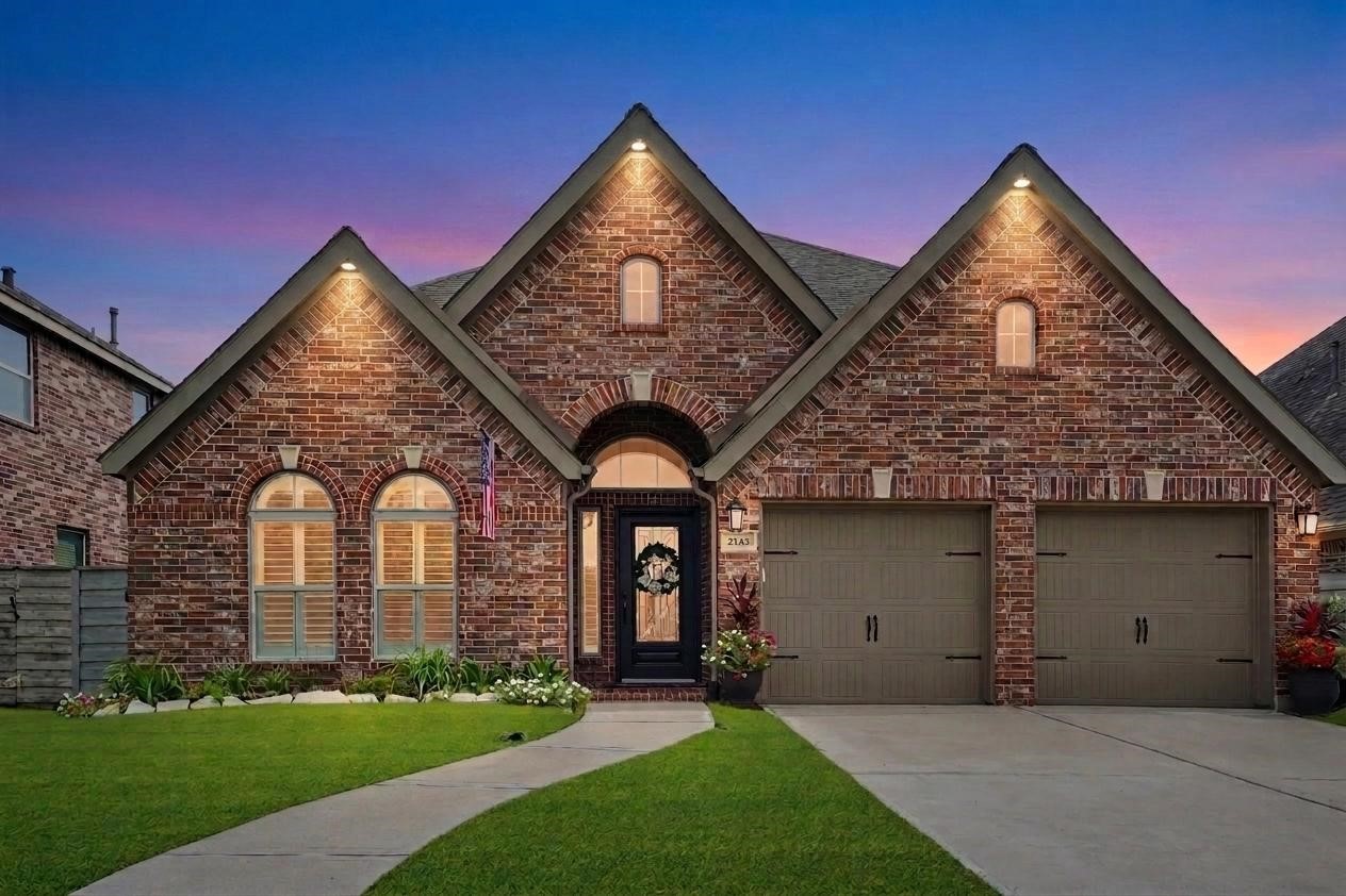 Charming brick home with a gabled roof, Exterior Lighting, double garage, and inviting front yard. The house features arched windows and a warmly lit entrance, perfect for a cozy, welcoming atmosphere.