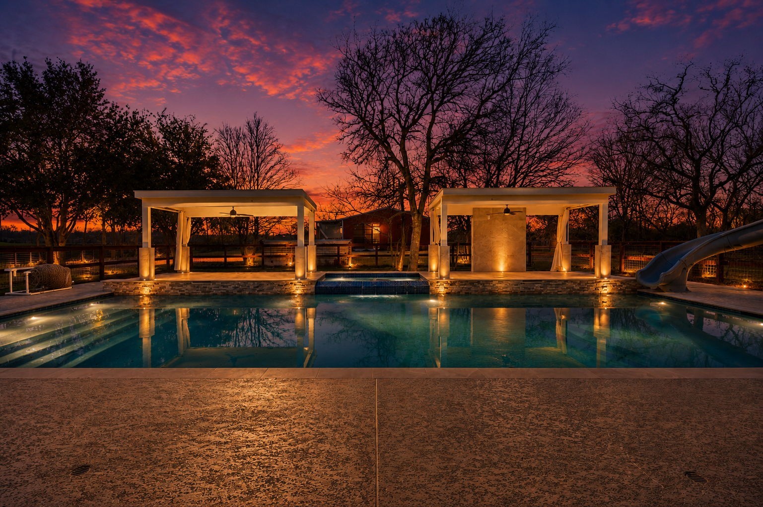 2730 Lawrence Road Kemah, TX 77565 - Photo 3 of 46 This photo showcases a luxurious backyard with a beautifully lit pool at sunset. It features two elegant cabanas, ambient lighting, and a slide, creating an inviting outdoor oasis perfect for relaxation and entertainment.
