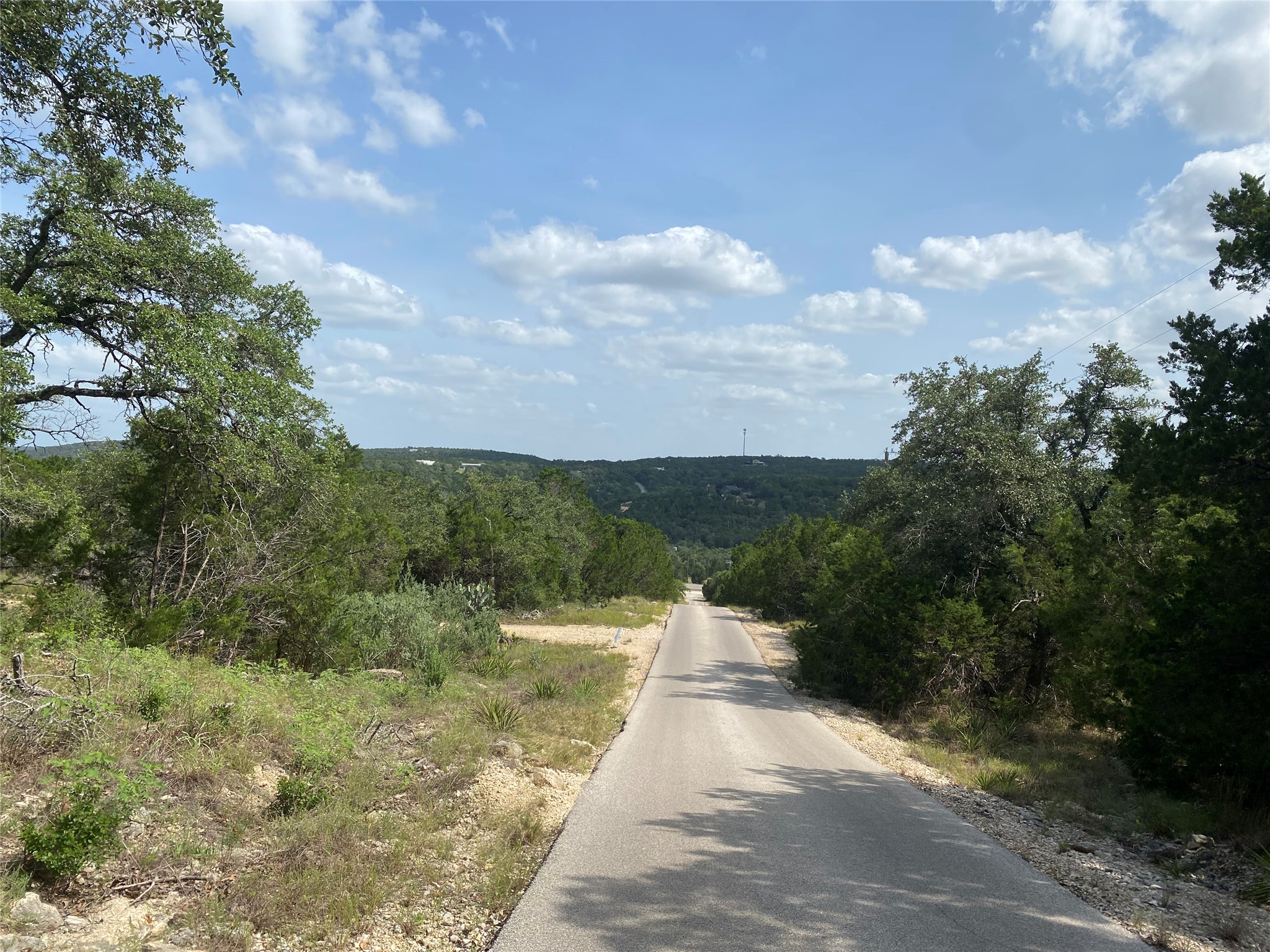 Tbd Whisper Ridge Wimberley, TX 78676 - Photo 5 of 7 An inviting paved road winds through a lush, tree-lined landscape under a vast blue sky.