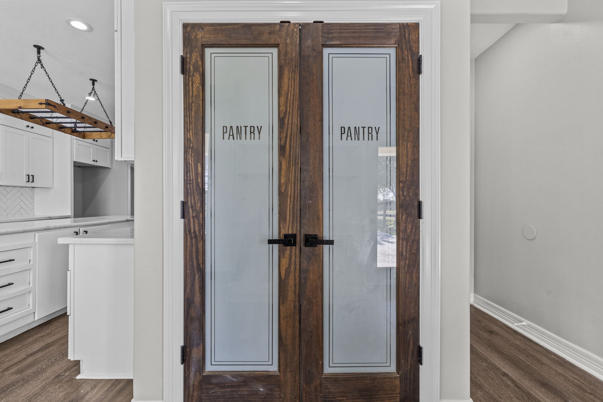 2730 Lawrence Road Kemah, TX 77565 - Photo 17 of 46 This photo showcases a modern kitchen with a stylish pantry featuring double doors with frosted glass panels and wooden frames. The kitchen has a sleek design with white cabinetry, a herringbone tile backsplash, and contemporary lighting fixtures. The flooring is a warm-toned wood, creating an inviting atmosphere.
