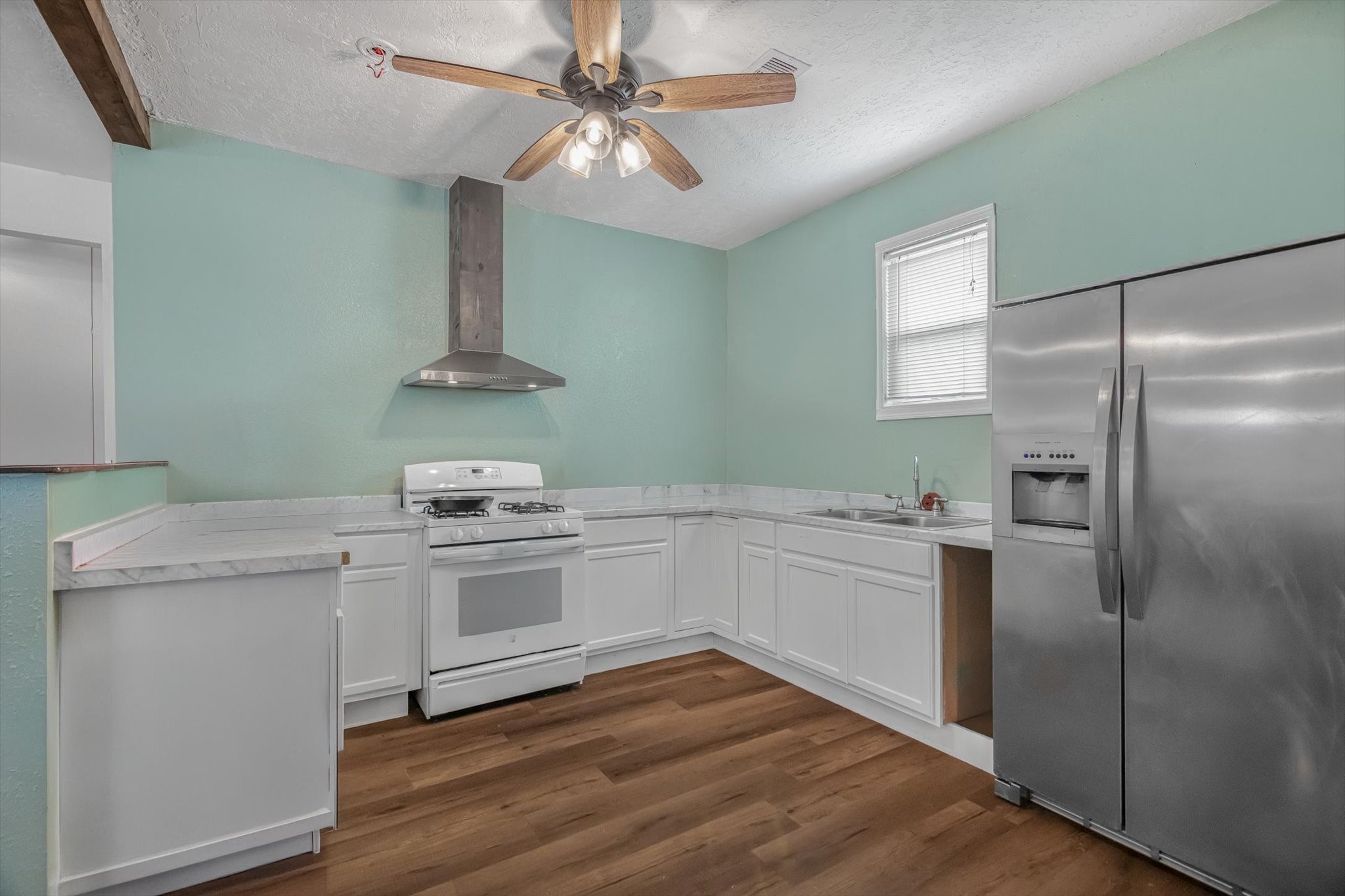 905 43rd Street Galveston, TX 77550 - Photo 34 of 43 Bright kitchen with mint green walls, featuring white cabinetry, a stainless steel fridge, and a gas stove. It has wood flooring, a ceiling fan, and ample countertop space with a modern exhaust hood.