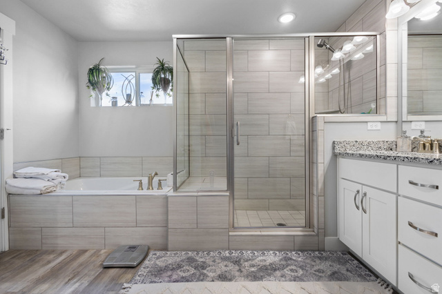 7889 Bristlecone Road Eagle Mountain, UT 84005 - Photo 25 of 42 Bathroom featuring wood-finish flooring, a tiled soaking tub with brass fixtures, a glass-enclosed shower, and a dual vanity with a speckled countertop and white cabinetry