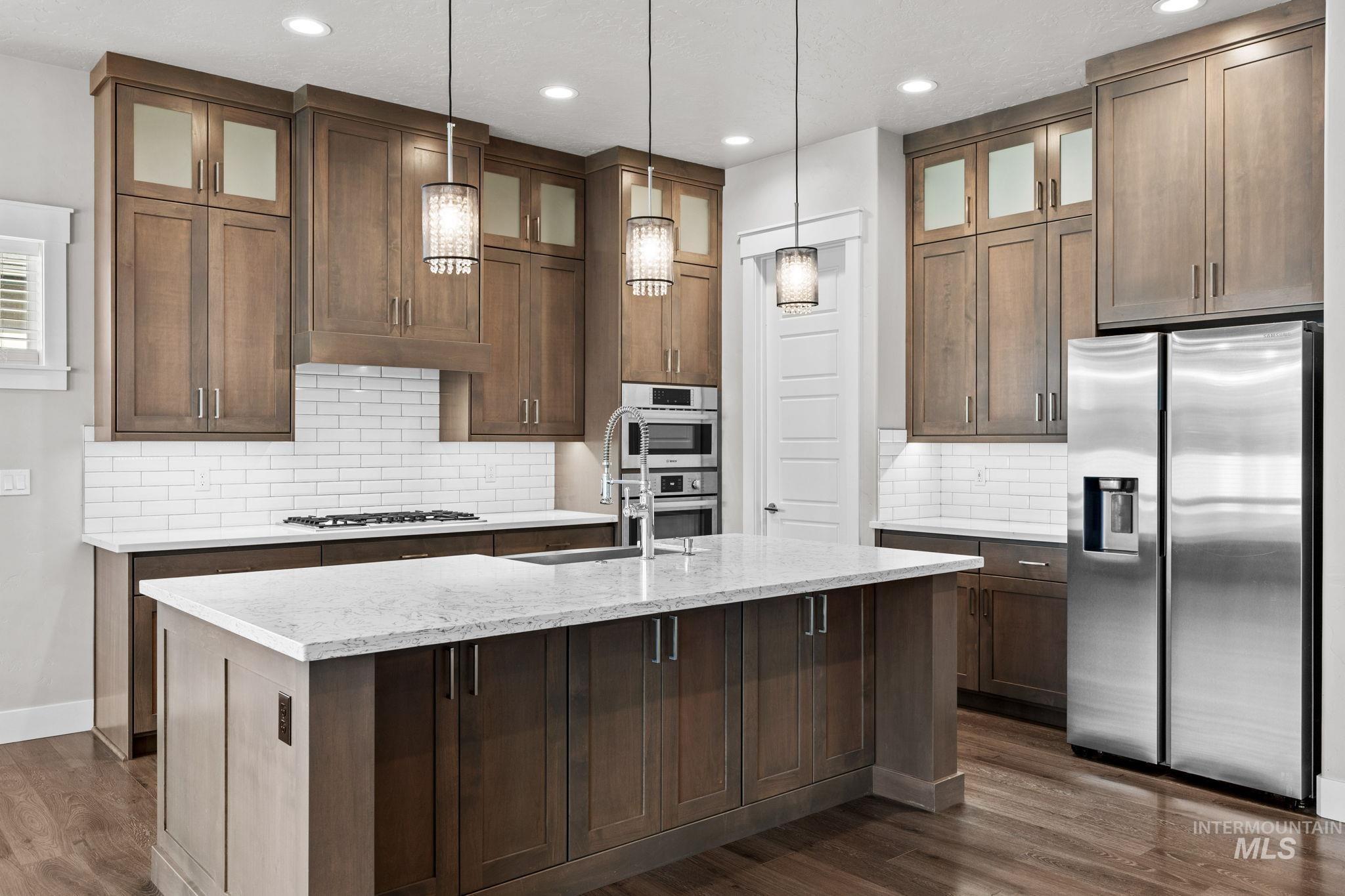 2934 East Murchison Street Meridian, ID 83642 - Photo 9 of 44 Open-concept kitchen featuring an oversized island with a light-toned countertop, wood-finish cabinetry, stainless steel appliances, a subway tile backsplash, and recessed and pendant lighting
