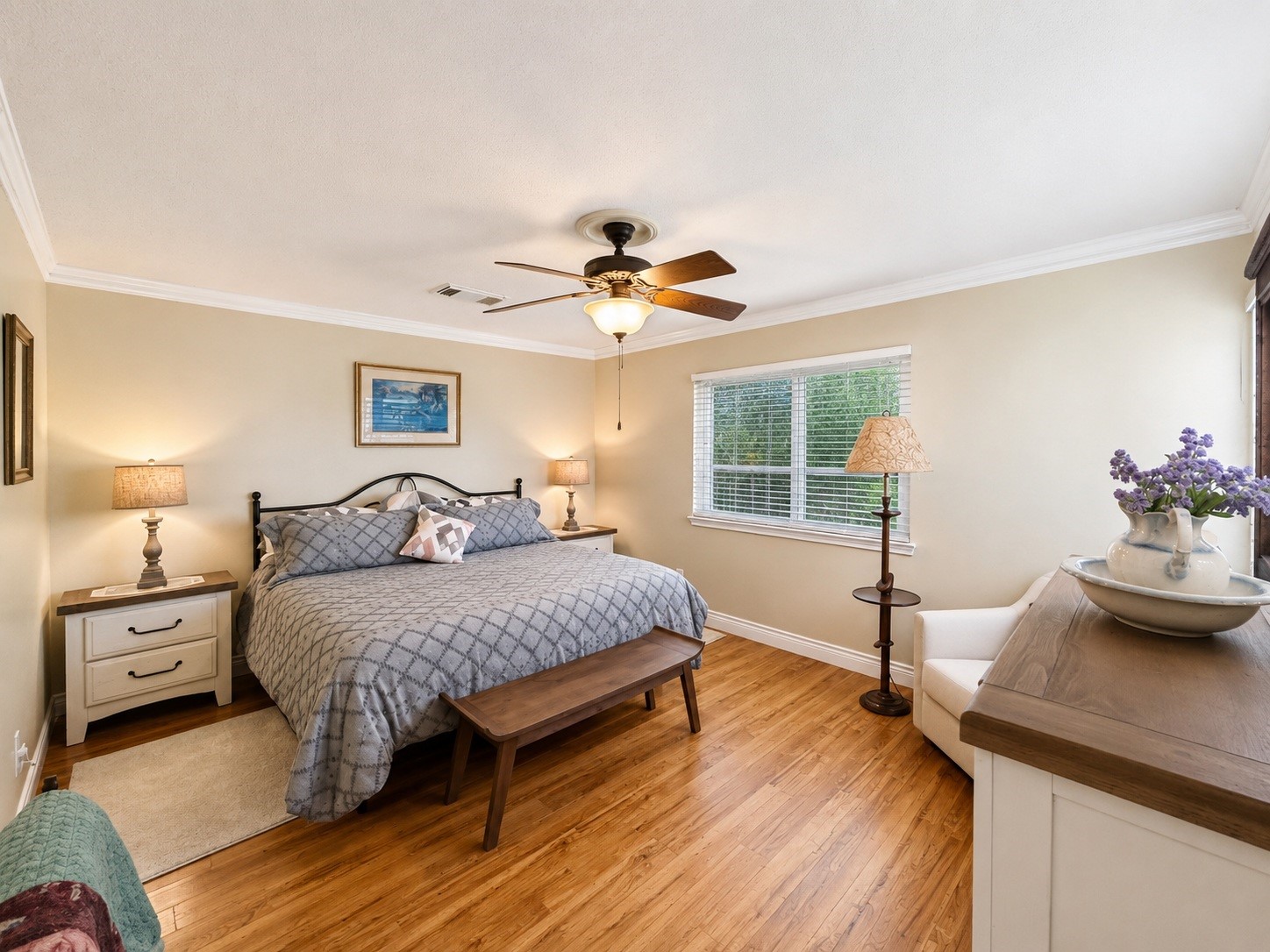 135 Sand Shoals Road Freeport, TX 77541 - Photo 12 of 20 Bright and inviting bedroom featuring hardwood floors, a ceiling fan, and ample natural light from a large window. The room is well-furnished with a comfortable bed, nightstands, and cozy decor, creating a warm and relaxing atmosphere.