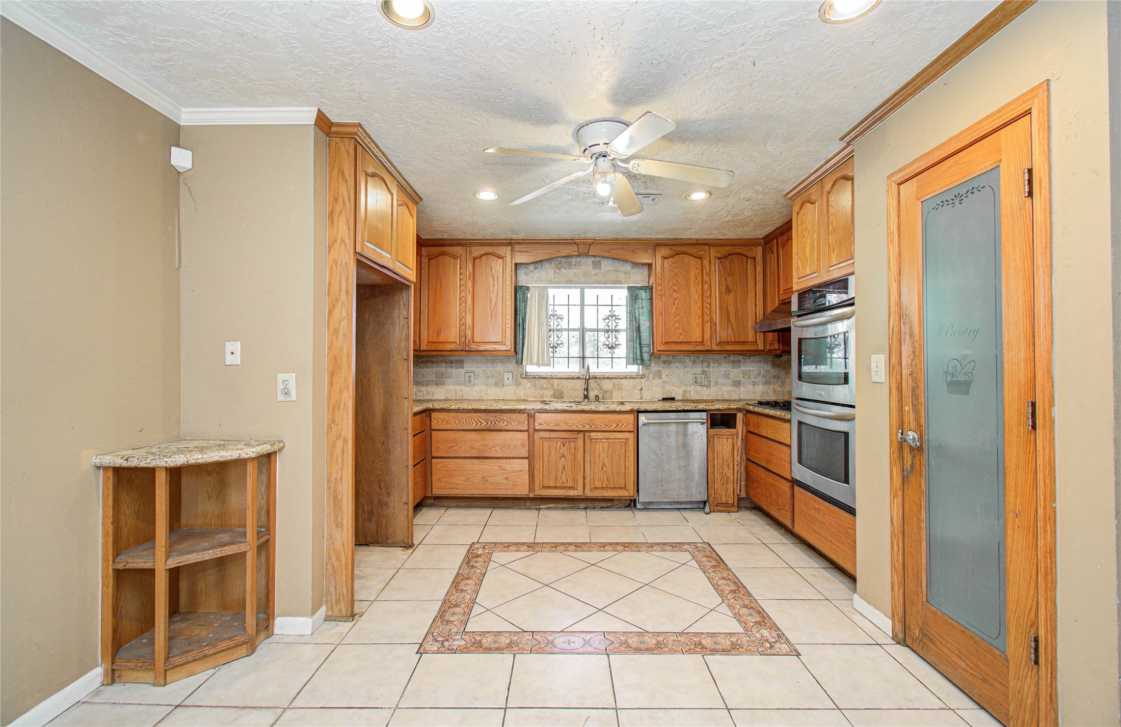 8428 Swiftwater Lane Houston, TX 77075 - Photo 13 of 26 This kitchen features warm wooden cabinetry, a double oven, and a dishwasher. It has a decorative tile floor, a ceiling fan, and a pantry with a frosted glass door. The space is bright with ample natural light from the window above the sink.