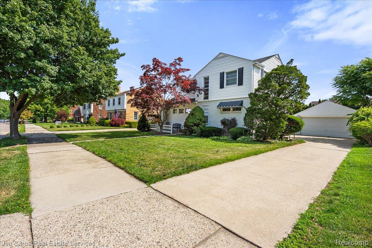 1010 Nottingham Road Grosse Pointe, MI 48230 - Photo 2 of 30 White two-story residence featuring black shutters and awnings, a well-maintained front lawn, mature landscaping, and a detached two-car garage with a concrete driveway