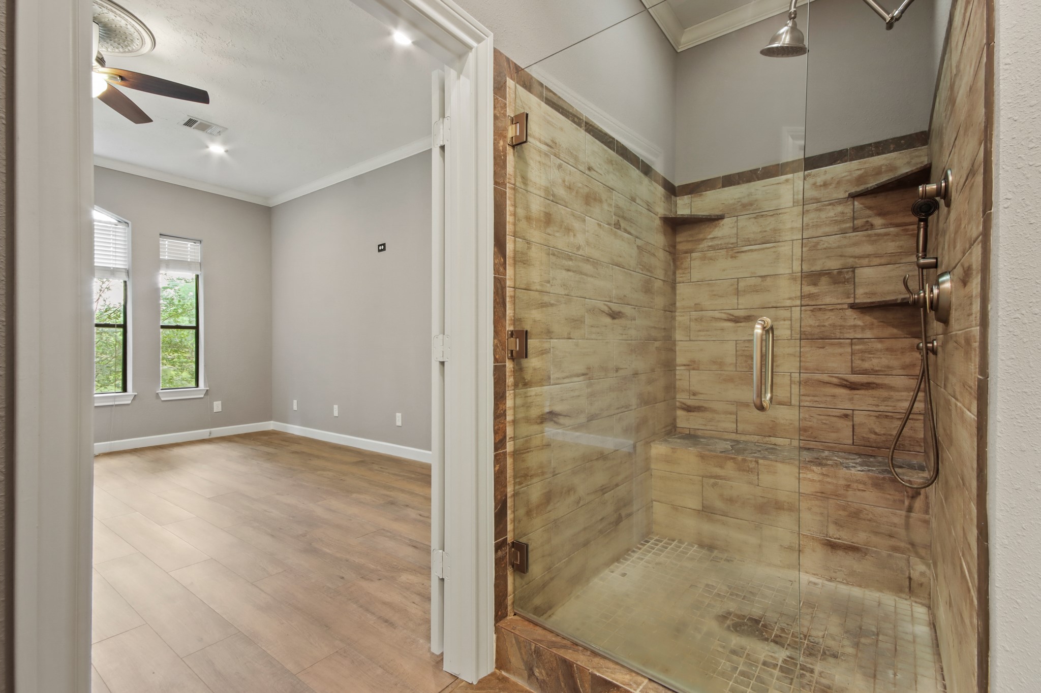 35 Susan Drive Cleveland, TX 77327 - Photo 12 of 33 This photo showcases a modern bathroom connected to a spacious bedroom. The bathroom features a glass-enclosed shower with wood-look tiles, while the bedroom has wood flooring and ceiling fan, offering a bright, airy feel with natural light from large windows.