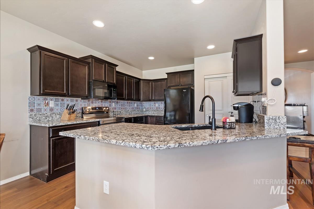 6308 South Kelso Way Boise, ID 83709 - Photo 10 of 35 Kitchen featuring a peninsula, dark wood finish cabinets, light stone counters, decorative backsplash, and black appliances