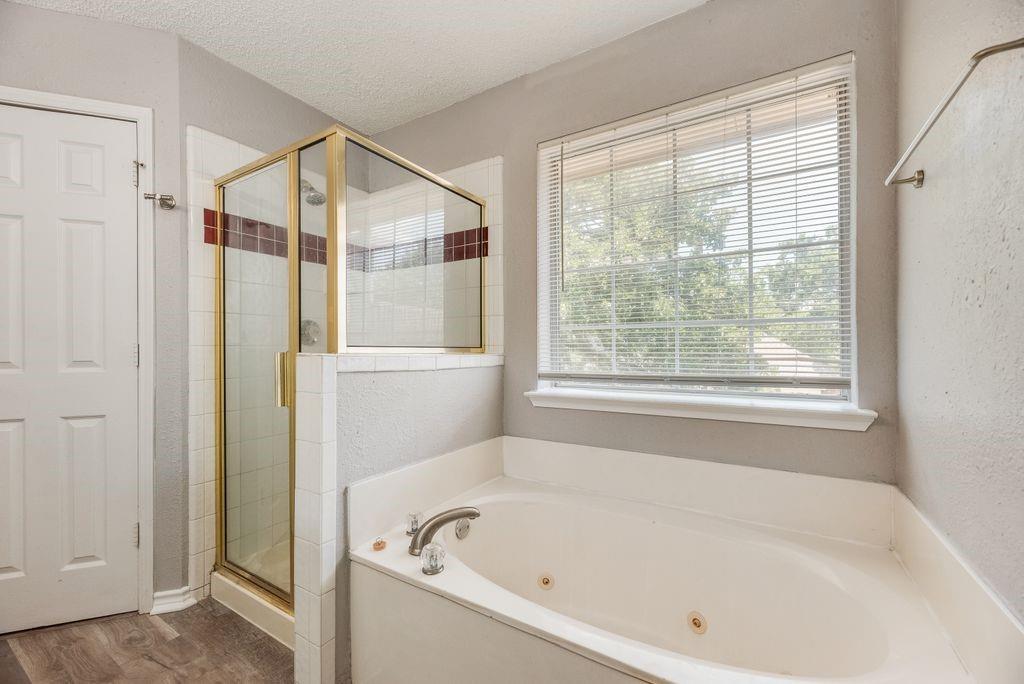 5610 Eagle Rock Road Arlington, TX 76017 - Photo 23 of 33 Spacious bathroom featuring a corner jetted soaking tub, separate glass-enclosed shower with tile surround, large window with blinds, and wood-finish flooring
