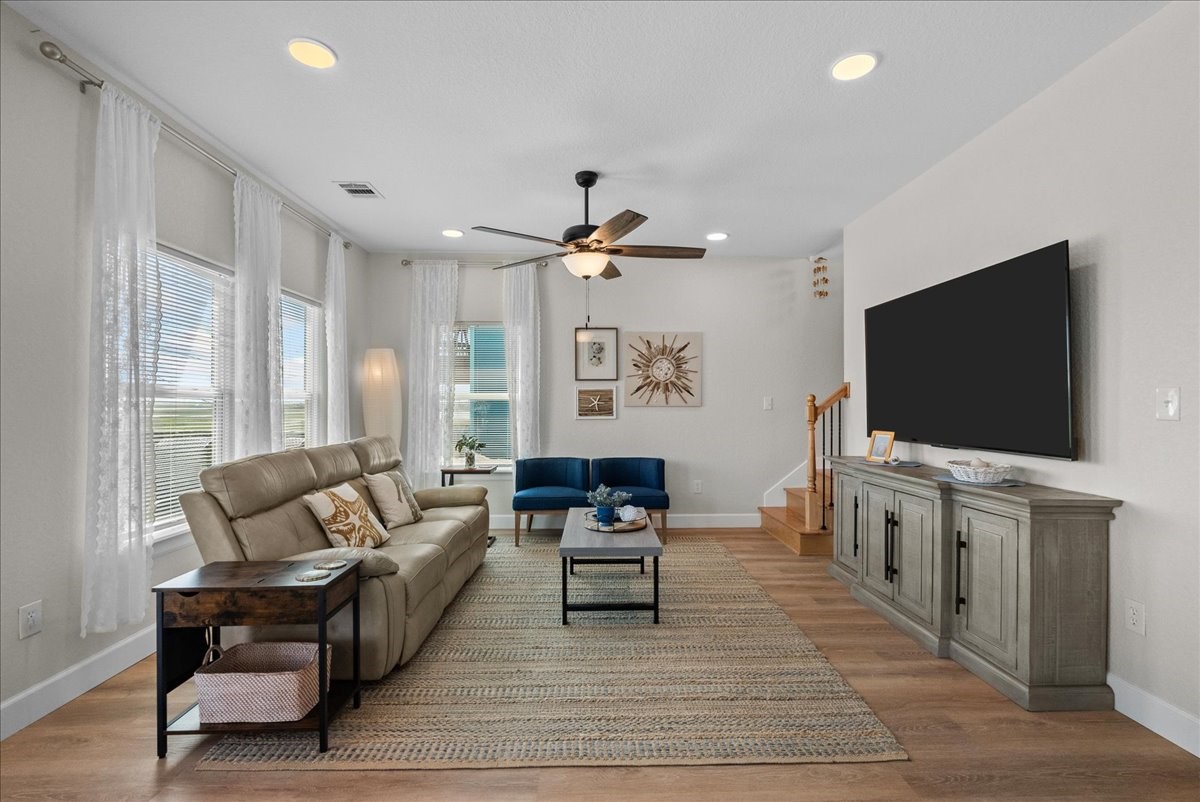 130 Beachcomber Avenue Surfside Beach, TX 77541 - Photo 14 of 33 Bright living room with large windows, neutral walls, and wood flooring. Features a comfortable sofa, blue accent chair, ceiling fan, and wall-mounted TV. Decor includes a stylish rug and modern artwork, with stairs leading up.