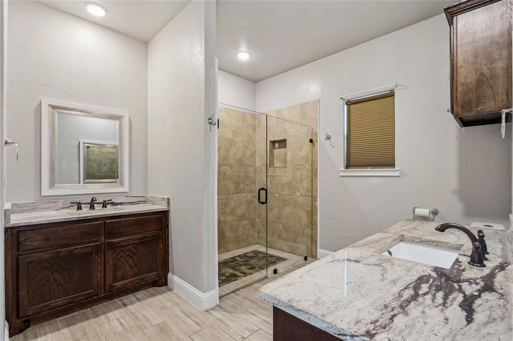 1502 Reed Street Greenville, TX 75401 - Photo 16 of 26 Bathroom featuring a glass-enclosed shower with tiled surround, wood-finish flooring, two distinct vanity areas with undermount sinks, and natural stone countertops