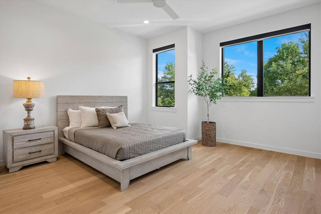 Bright primary bedroom showcasing wood-finish flooring, expansive black-framed windows with natural light, a gray upholstered bed frame with coordinating nightstand featuring drawer storage, and a potted accent plant