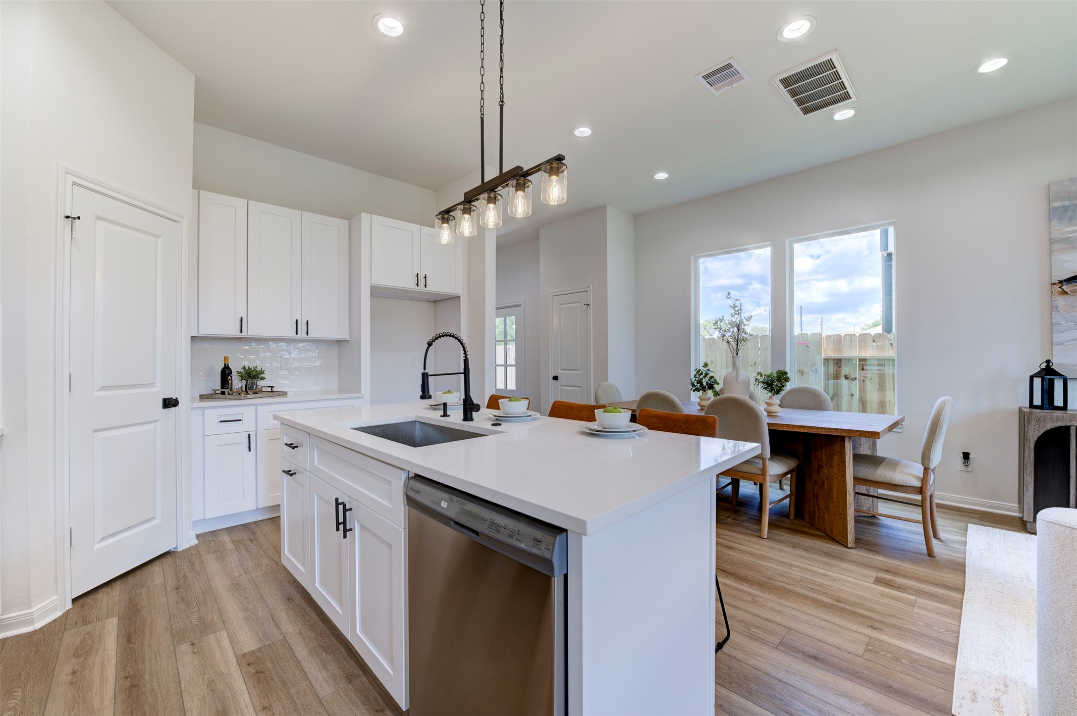 6807 Paris Street Houston, TX 77021 - Photo 16 of 50 Kitchen island with quartz countertop, farmhouse sink, matte black coil-spring faucet, stainless dishwasher, and Edison rail pendant overhead. Pantry door and appliance niche visible on left. Open sightline to dining area and large rear windows with backyard view.