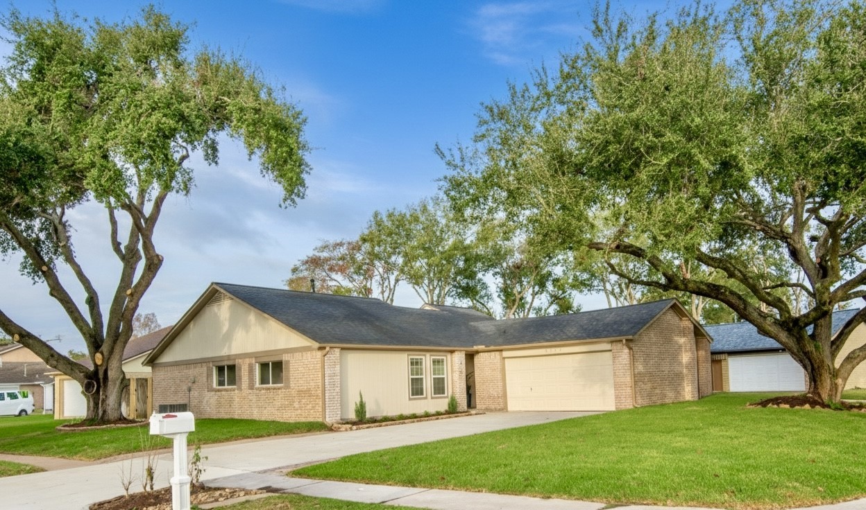 Single-story brick home with a two-car garage. The house is surrounded by a well-maintained lawn and mature trees, providing shade and curb appeal. The driveway and walkway lead to the front entrance, creating an inviting look.